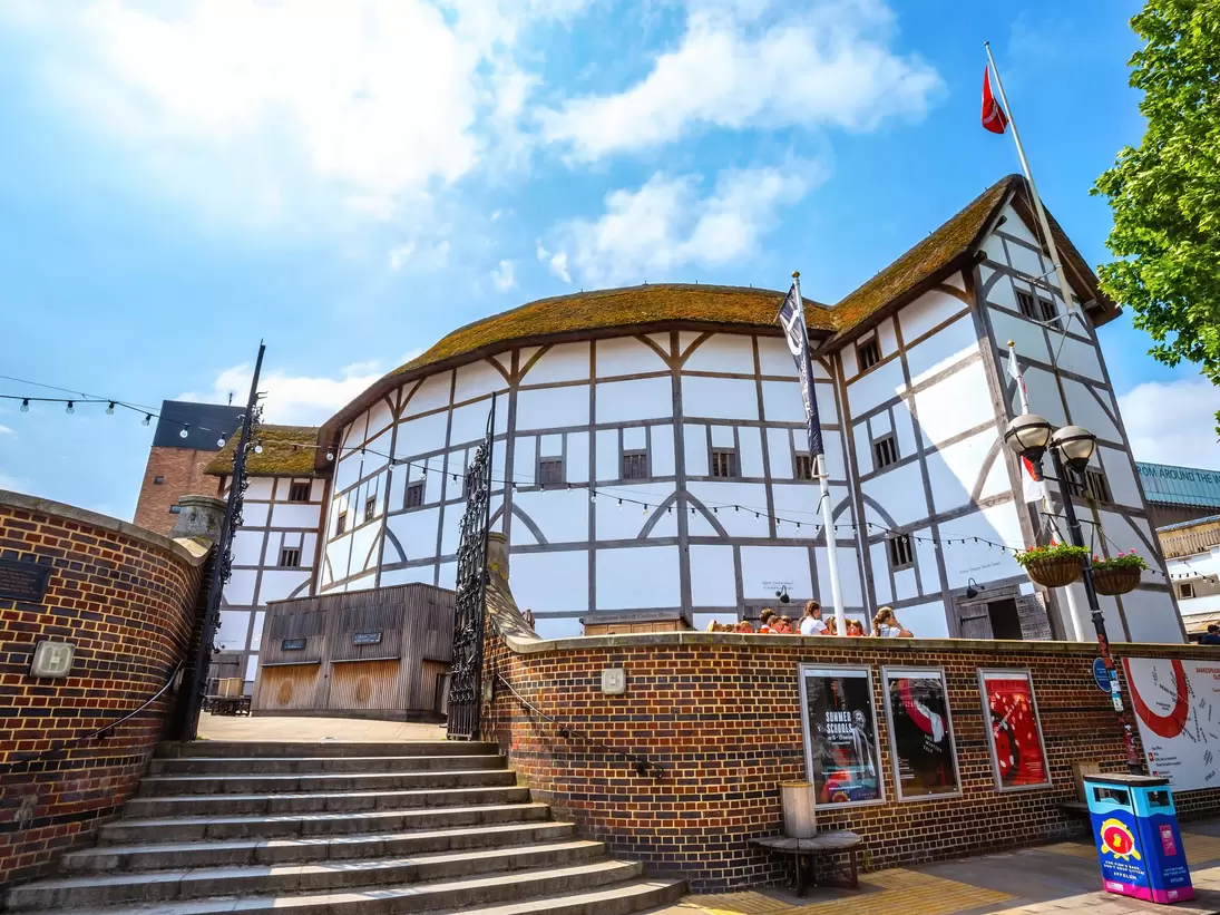 White half-timbered round playhouse under a thatched roof, above a curved brick wall, steps, flags and posters.