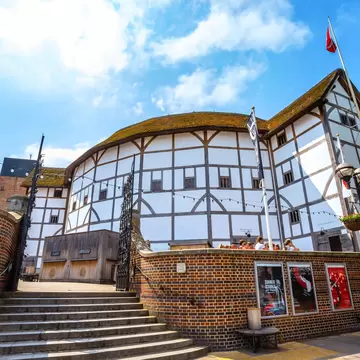 White half-timbered round playhouse under a thatched roof, above a curved brick wall, steps, flags and posters.