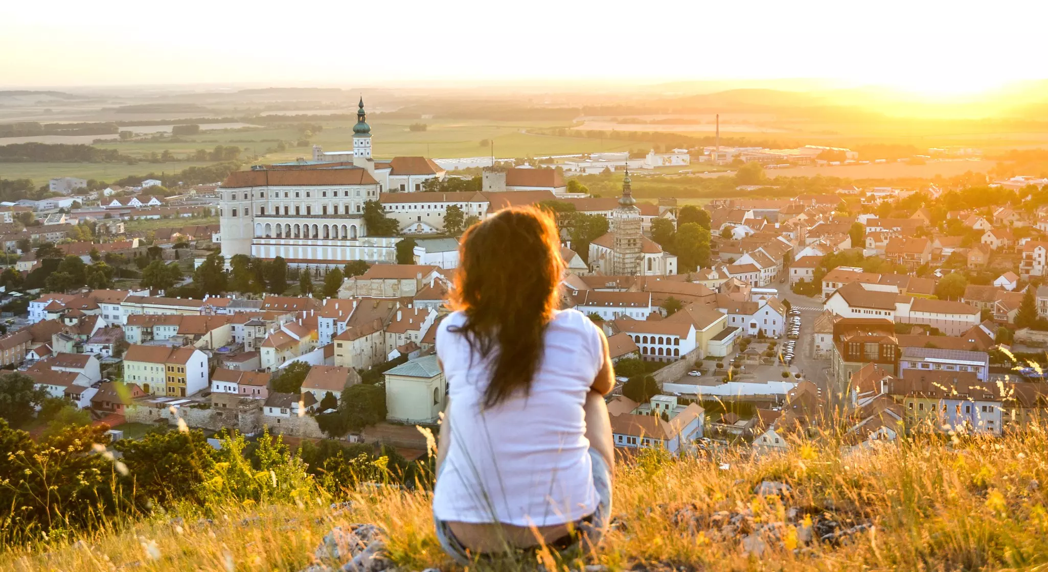 View of sitting young tourist woman overlooking beautiful Mikulov castle from the top of Saint Hill while sunset. South Moravia, wine region in the Czech Republic.