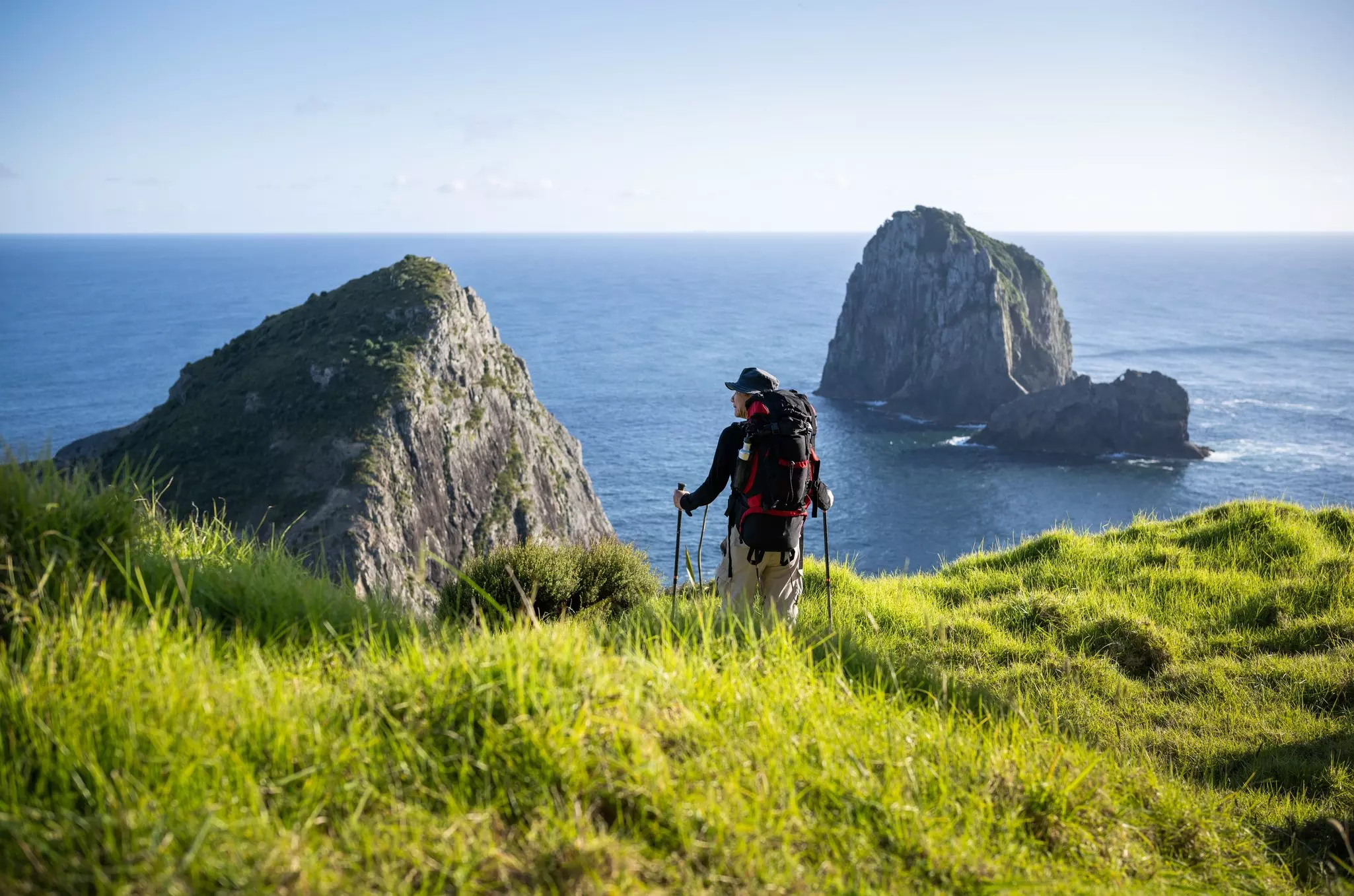 A hiker wearing a backpack and holding two hiking poles walks through grass on a cliff above the sea; there are two large rock formations in the blue water.