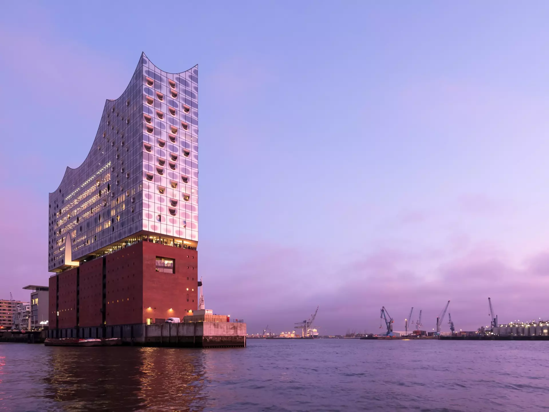 The Elbphilharmonie, viewed from the river, at dusk. A pink ligh is reflecting on the building's mirrored surface