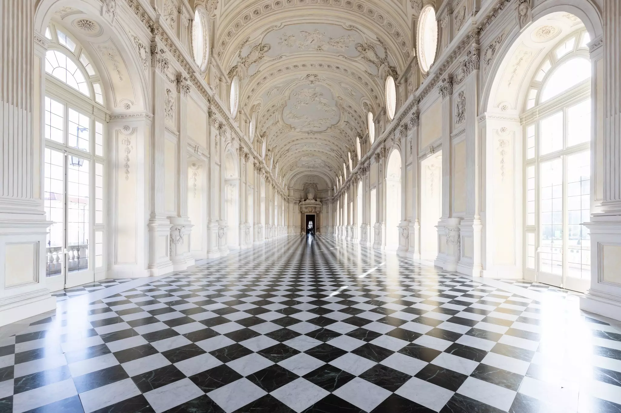 A vaulted gallery with ornate decoration is pictured. The windows and black-and-white-tiles render the room in linear perspective.