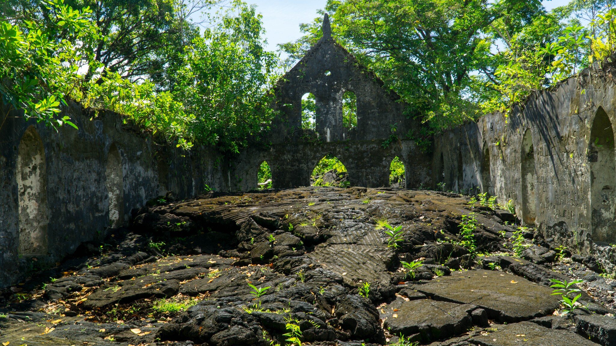 Moss-streaked stone walls and an arched gable enclosing a floor of ridged black lava, trees growing over the walls