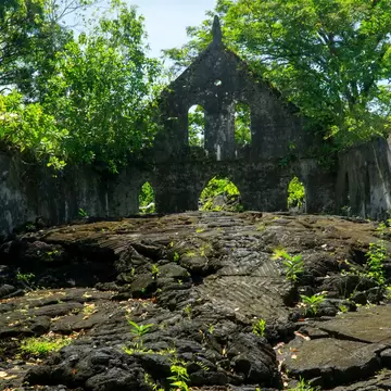 Moss-streaked stone walls and an arched gable enclosing a floor of ridged black lava, trees growing over the walls