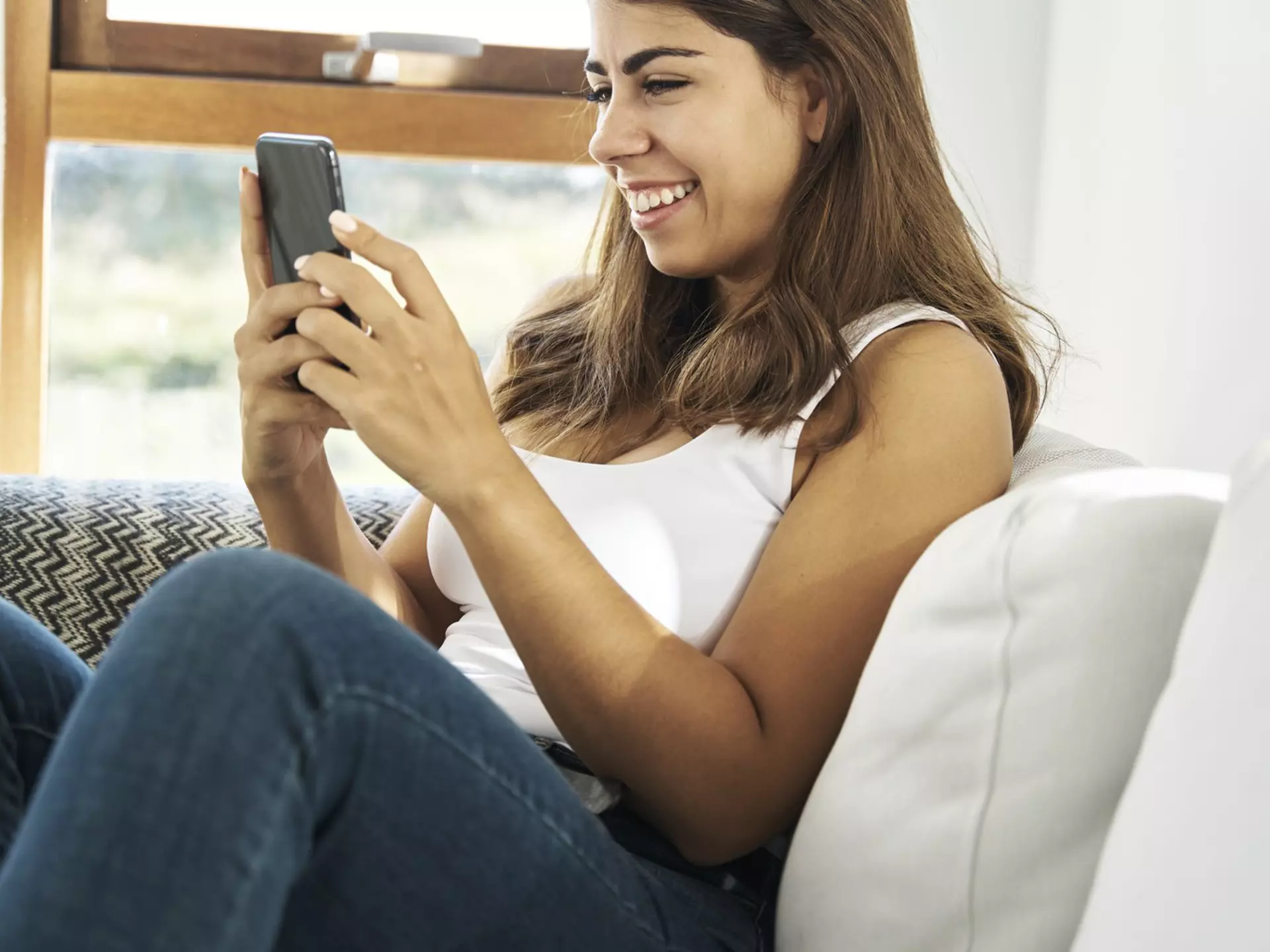 Cheerful young ethnic female in casual clothes sitting on cozy sofa near window and browsing mobile phone while resting at home