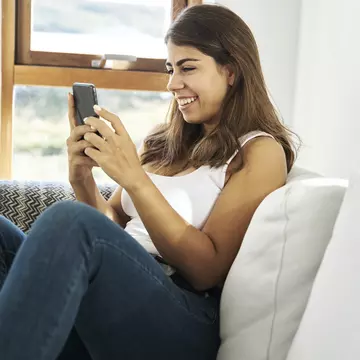 Cheerful young ethnic female in casual clothes sitting on cozy sofa near window and browsing mobile phone while resting at home
