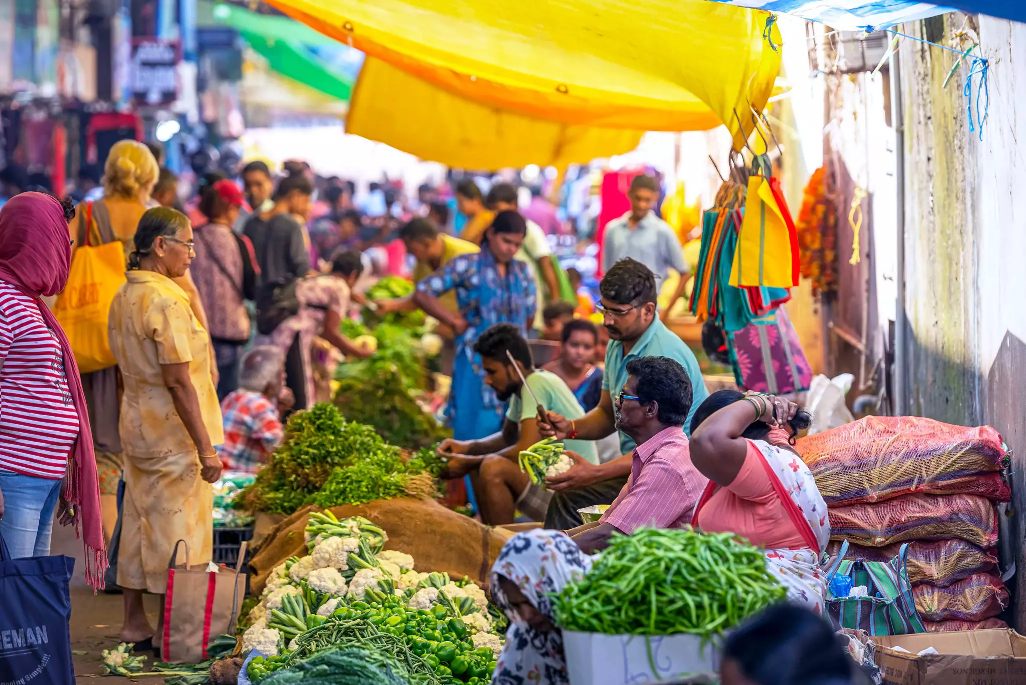 A busy market with people browsing and buying from stalls selling fresh vegetables.