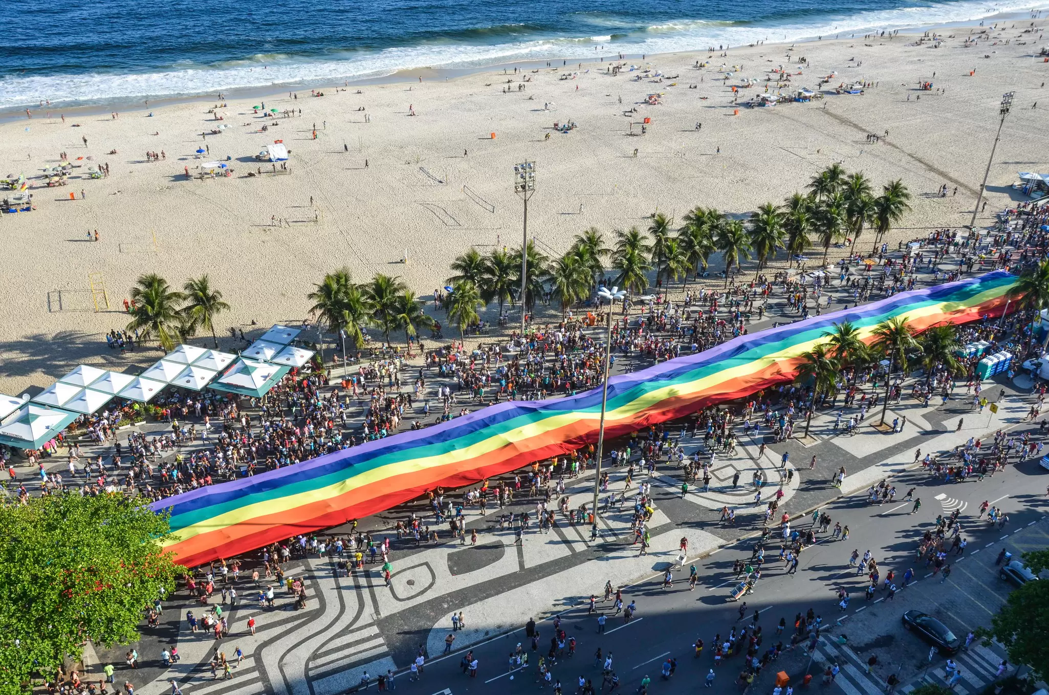 People along a beach carry long rainbow flag.