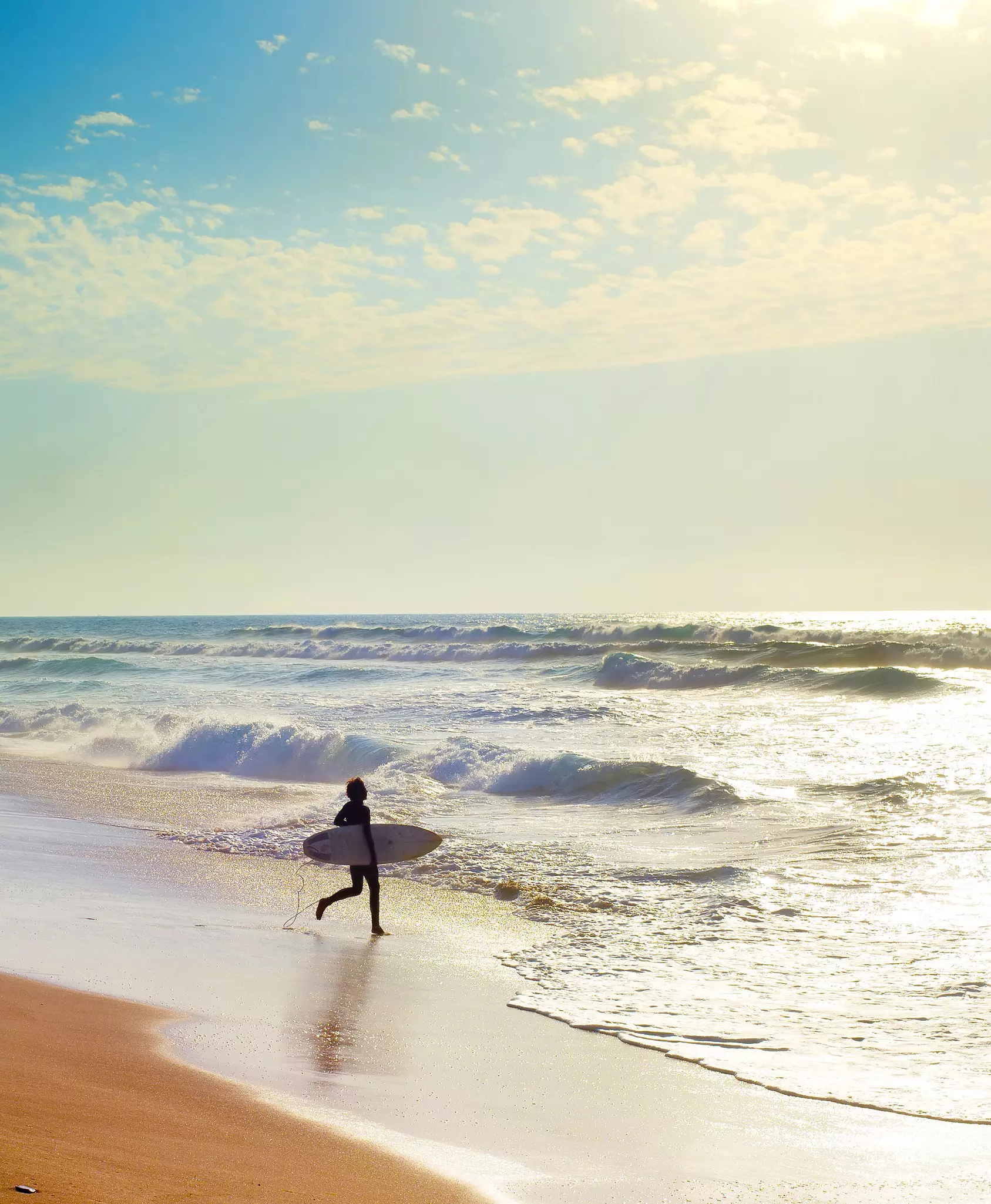 A surfer walks down the beach towards the sea in the blazing sunshine