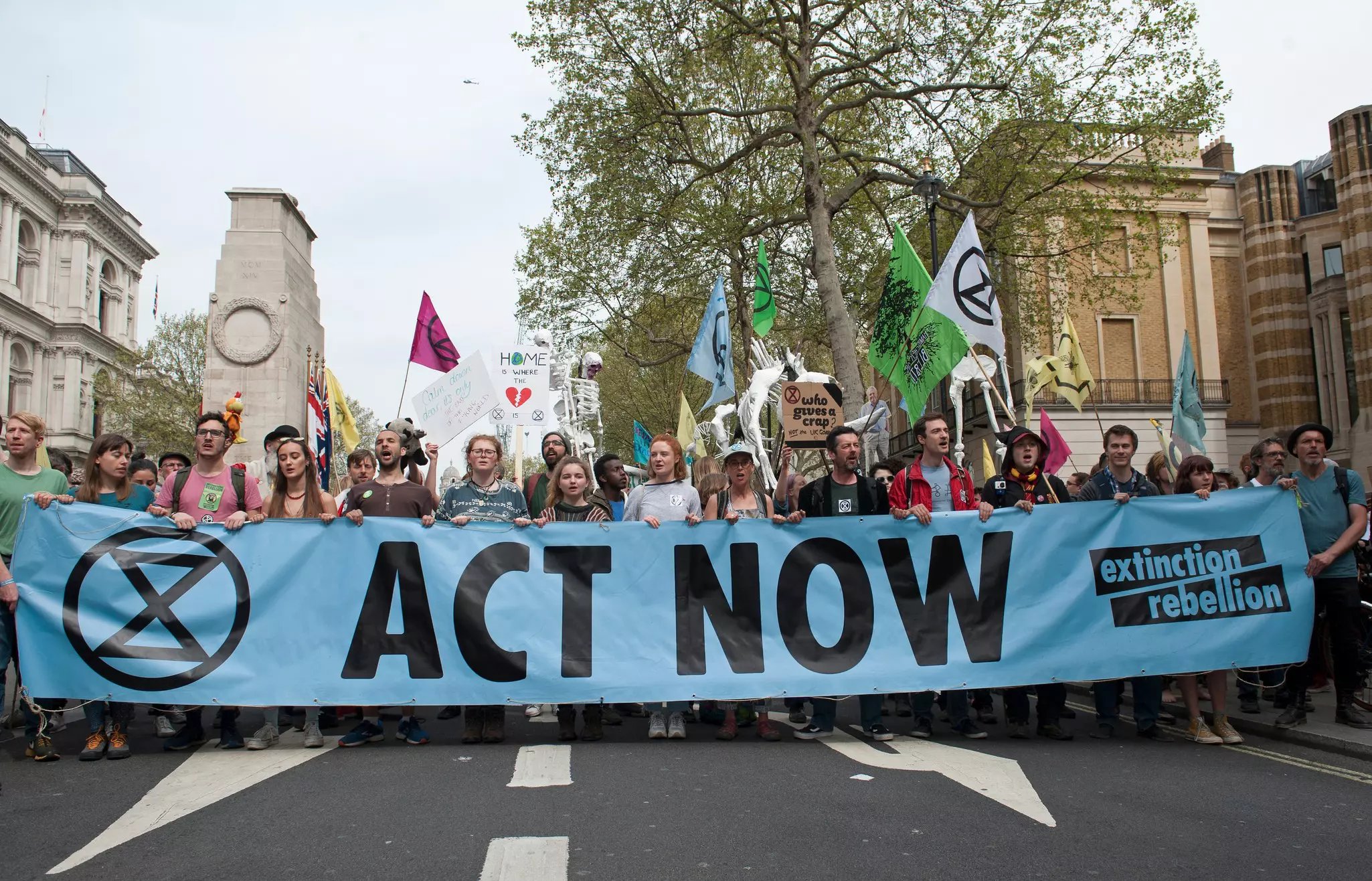 An image of Extinction Rebellion protested in central London. The crowd are carrying a large blue banner reading 'ACT NOW' as well as flags and other smaller signs.