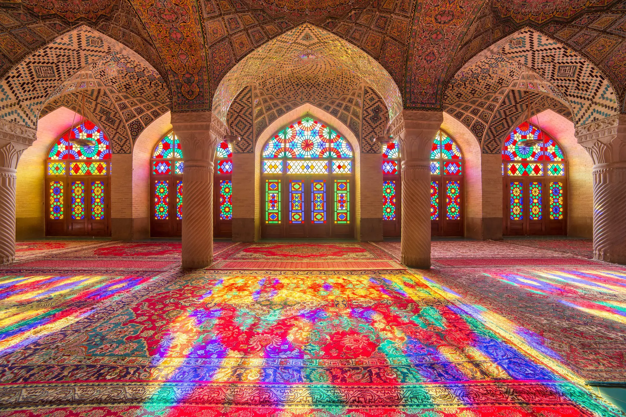 Light floods through stained-glass windows in a mosque casting colors all over the intricately patterened carpets.