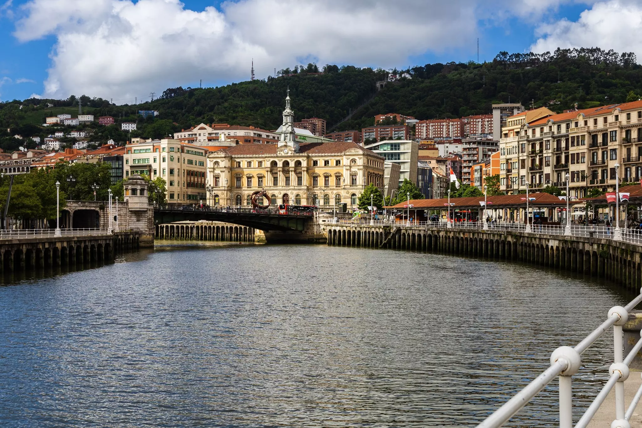 A river winds through the center of a city, with a grand building along the bank, and green hills rising above.