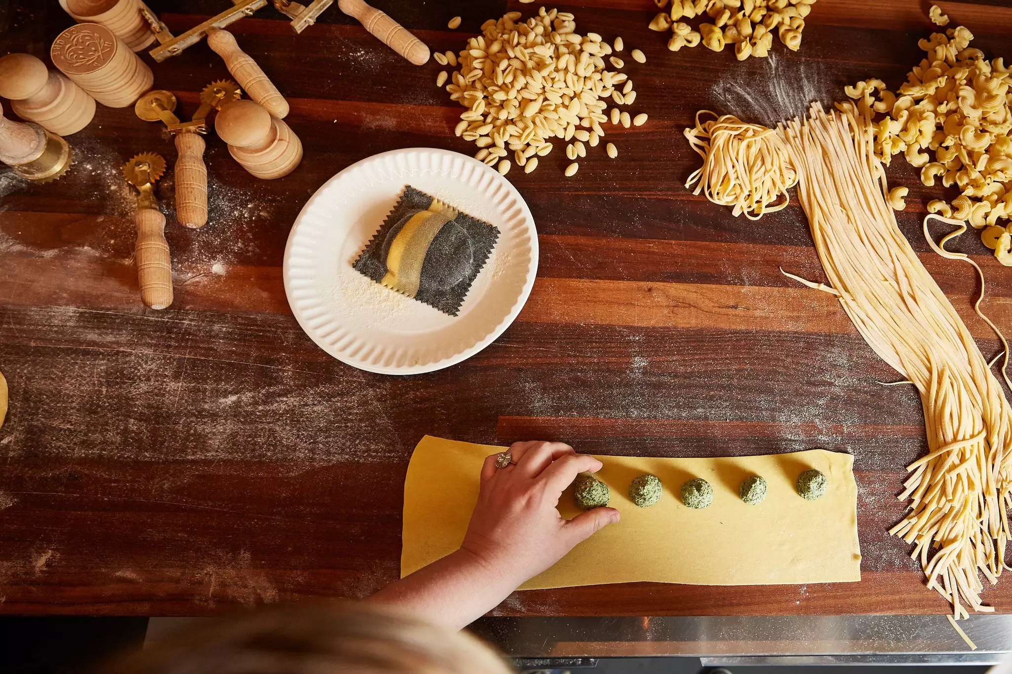 A hand placing round balls of filling on a sheet of pasta rolled out on a dark wood table; there are other pasta shapes on the table.