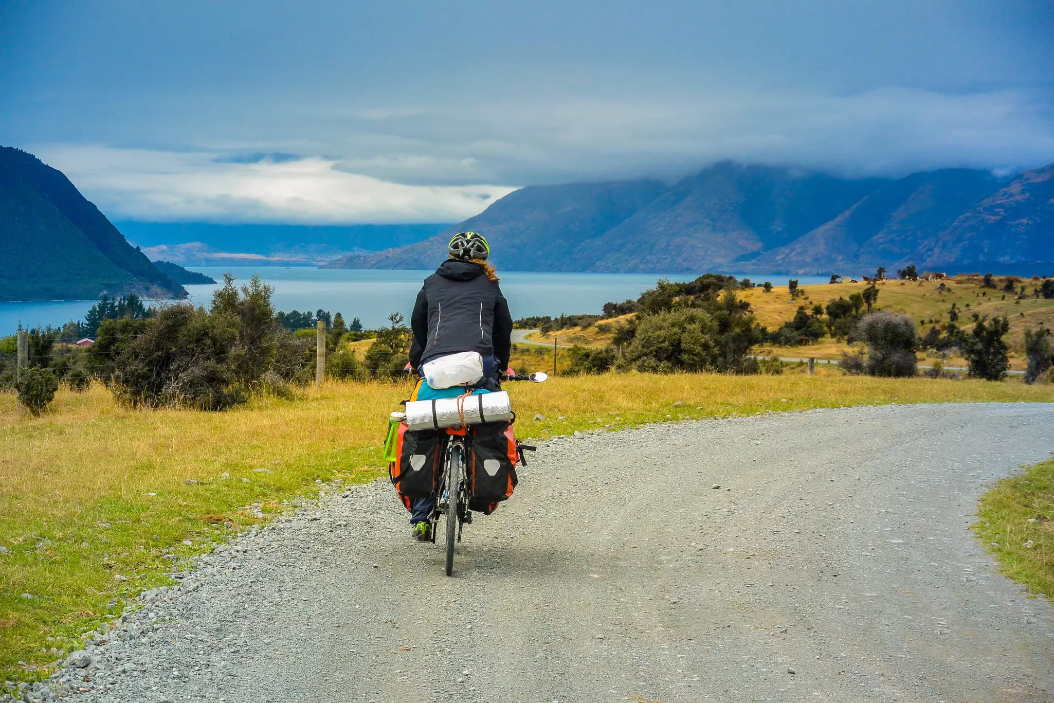 A cyclist riding a bike loaded with gear and supplies pedals along a gravel track towards a lake.