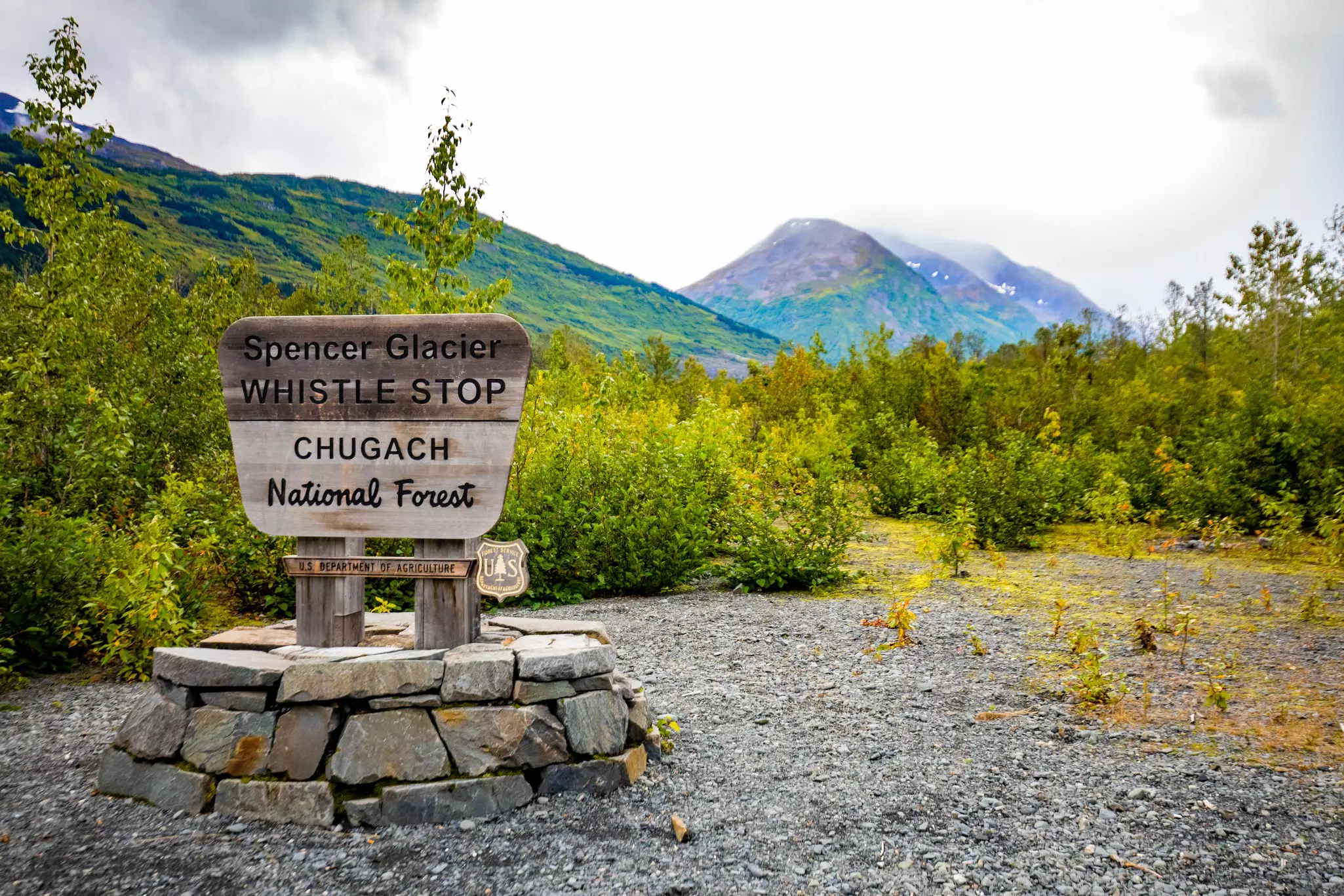 Wooden sign with the words "Spencer Glacier Whistlestop: Chugach National Forest" on a gravel area with greenery and mountains in the distance on a cloudy day.