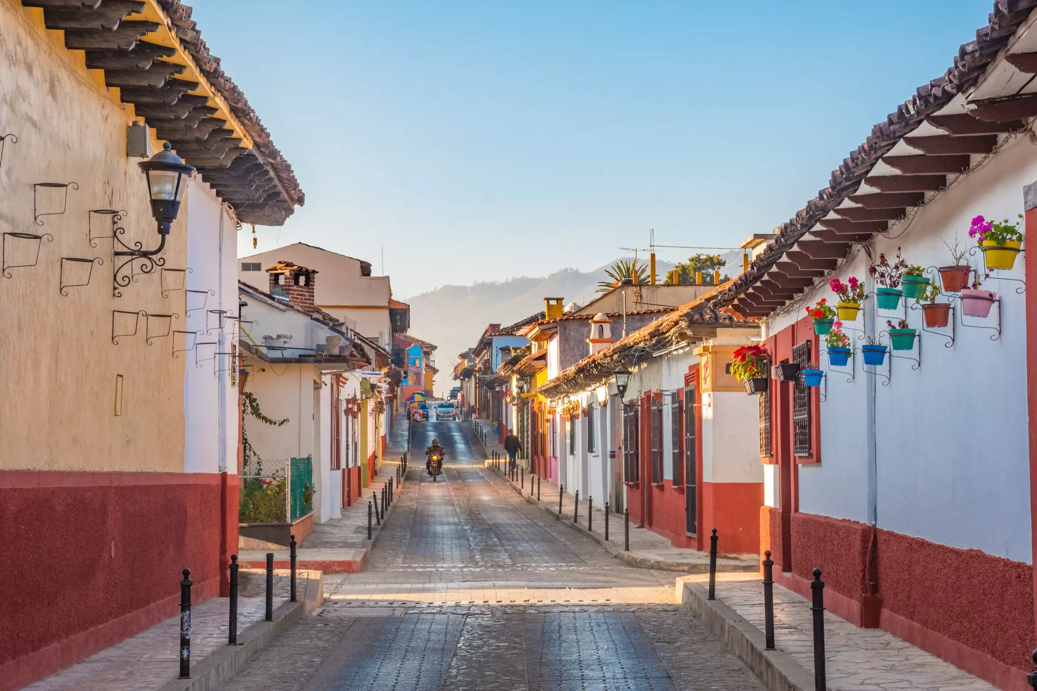 the sunlit streets of a colonial Mexican village. White buildings with red bottoms line a cobblestone street. A motorcyclist rides down the street.