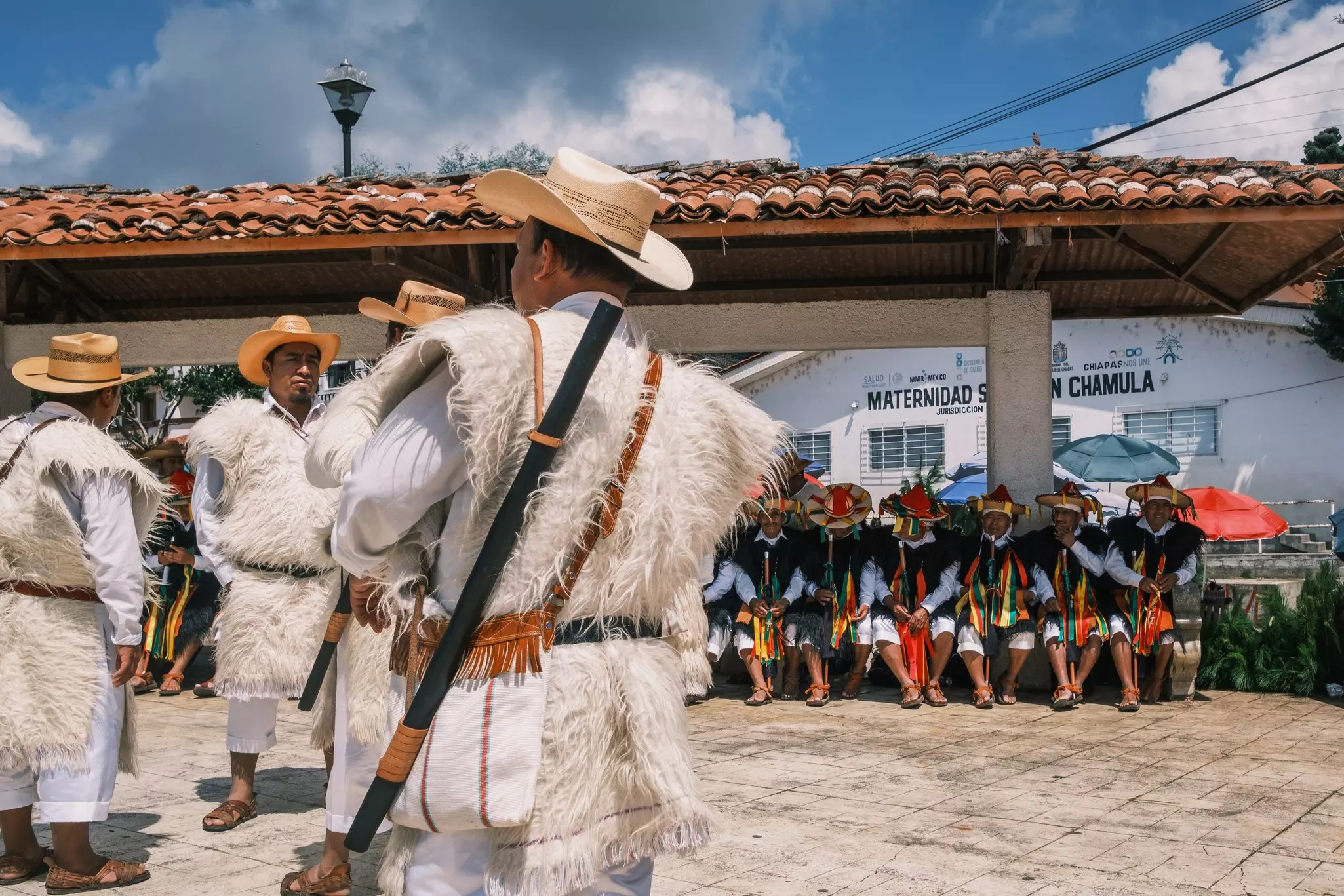Men in tunics made of animal skins wait at a city plaza, while others in traditional dress sit on a bench in the share.