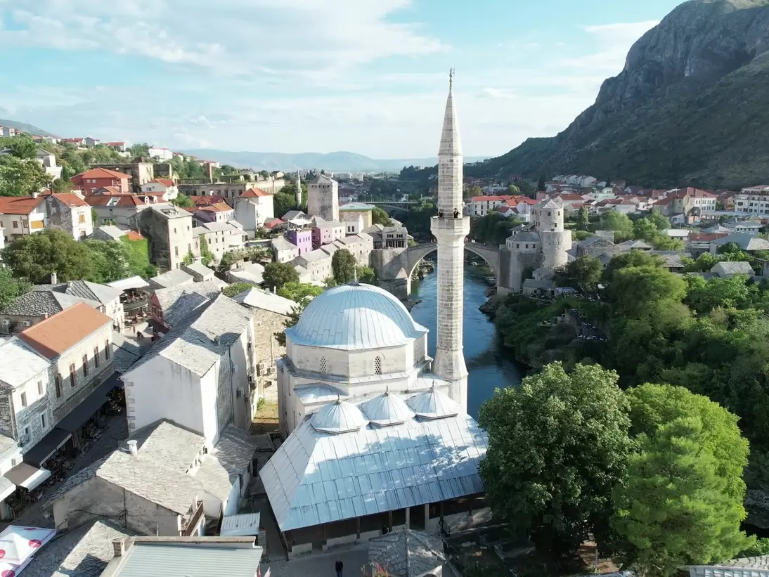 Domed Ottoman mosque with a tall stone minaret beside the Neretva River, old bridge and old town beyond, Mostar