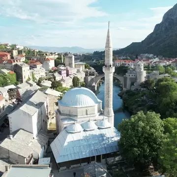 Domed Ottoman mosque with a tall stone minaret beside the Neretva River, old bridge and old town beyond, Mostar
