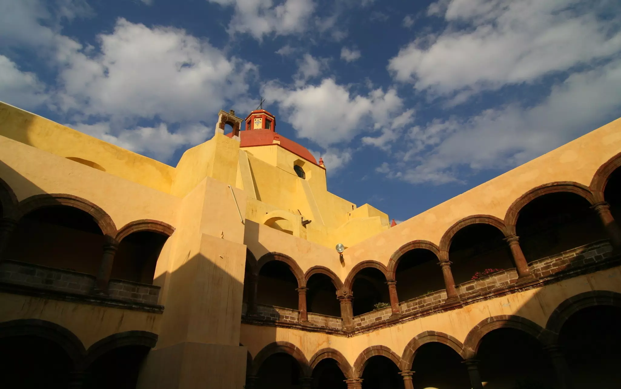 Close-up of yellow church building with smooth cement facade with a red roof and arched open windows along upper and lower walkways, with a light-blue sky and some clouds beyond.