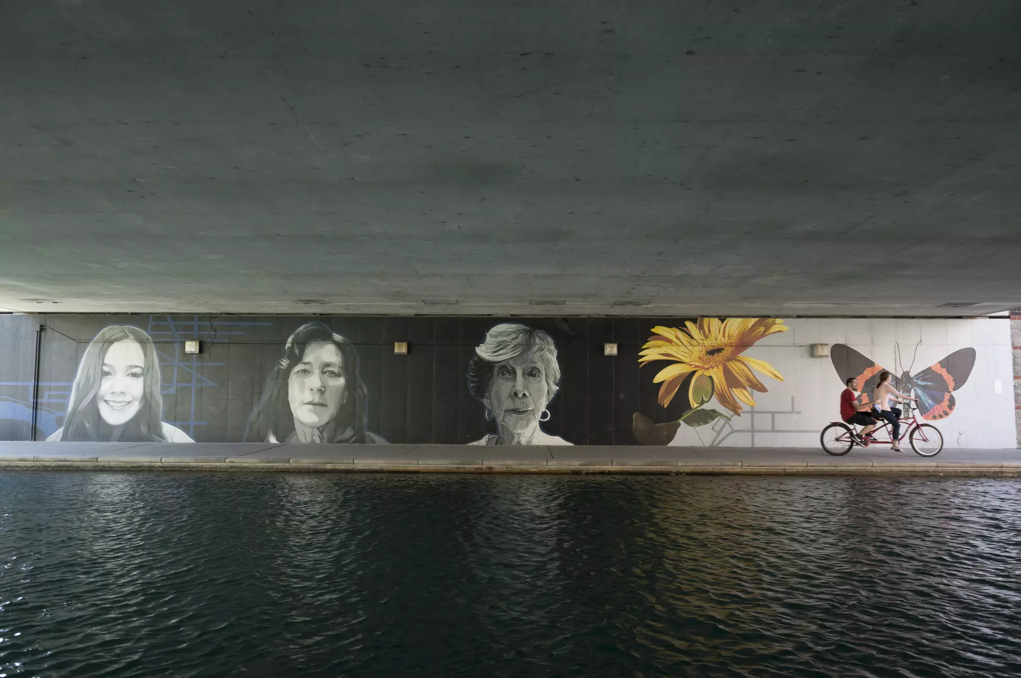 People on a tandem bike riding next to a body of water by a mural of three faces and a yellow flower