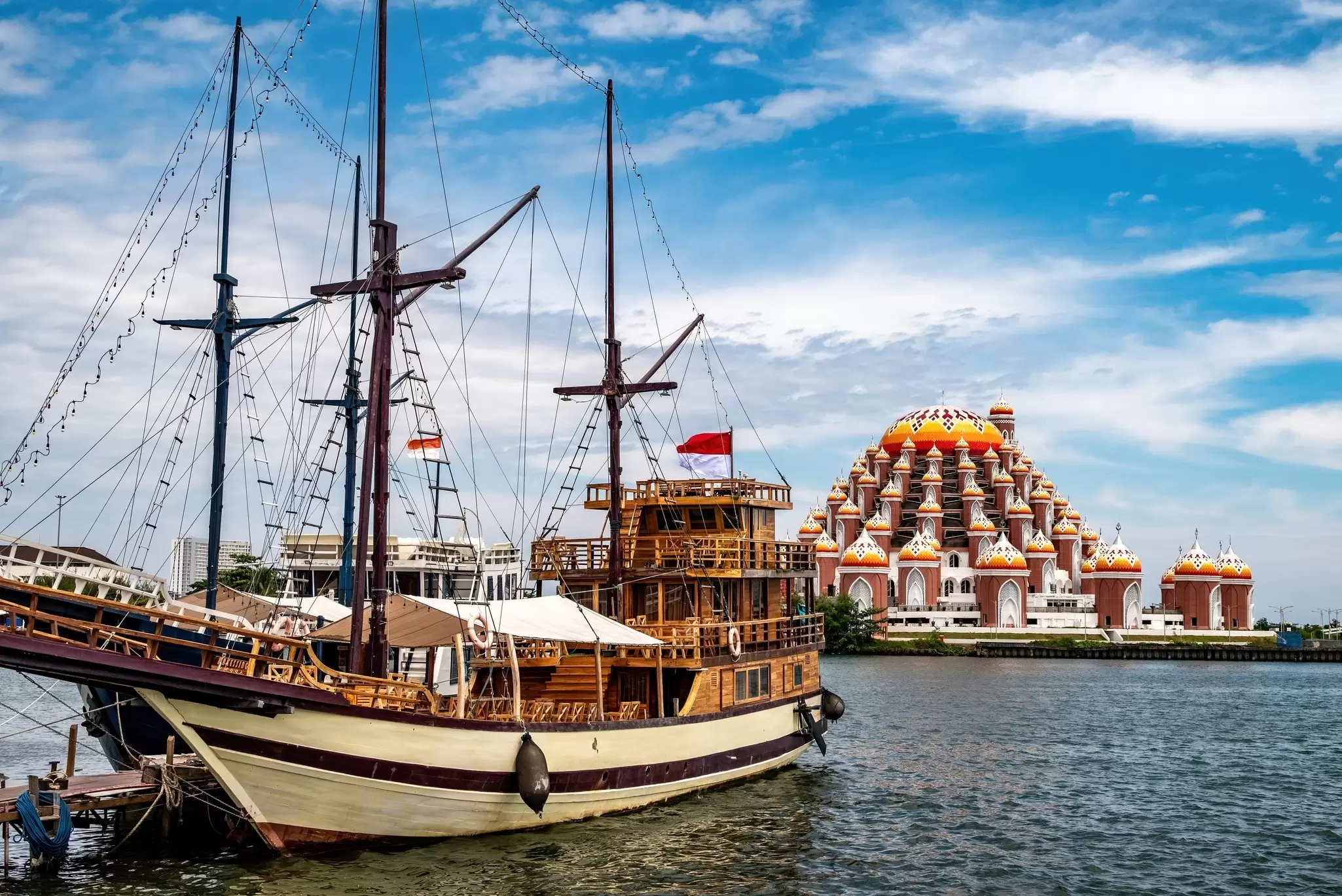 A traditional two-masted schooner is in port, with a terraced mosque visible on the far shore of the water, Makassaar, Sulawesi, Indonesia