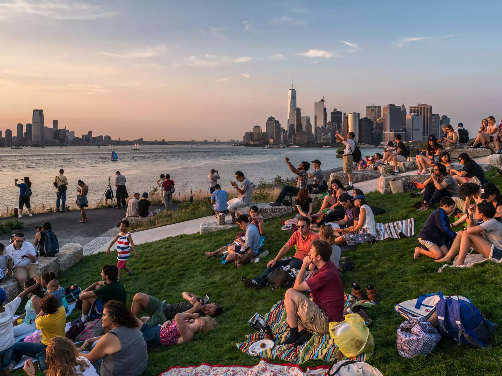 A crowd of people sitting on blankets watch the sunset from Governors Island with the New York City skyline in the background
