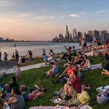 A crowd of people sitting on blankets watch the sunset from Governors Island with the New York City skyline in the background