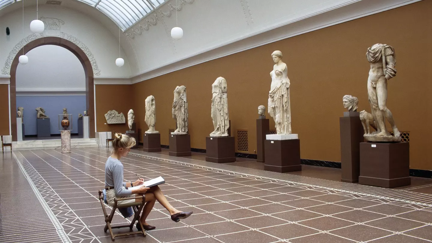 A blonde woman sits in front of classic sculptures while she sketches them in the Glyptotek in Copenhagen