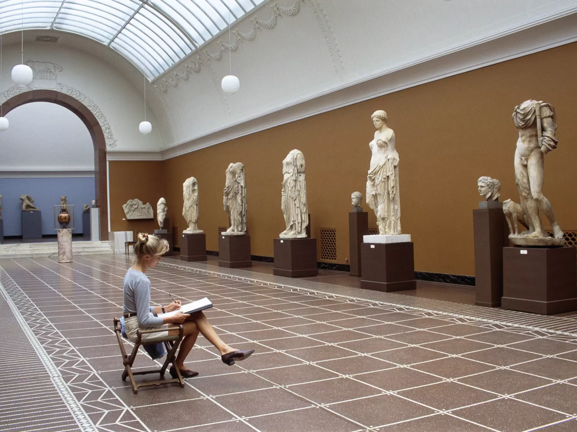 A blonde woman sits in front of classic sculptures while she sketches them in the Glyptotek in Copenhagen