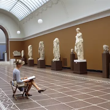 A blonde woman sits in front of classic sculptures while she sketches them in the Glyptotek in Copenhagen
