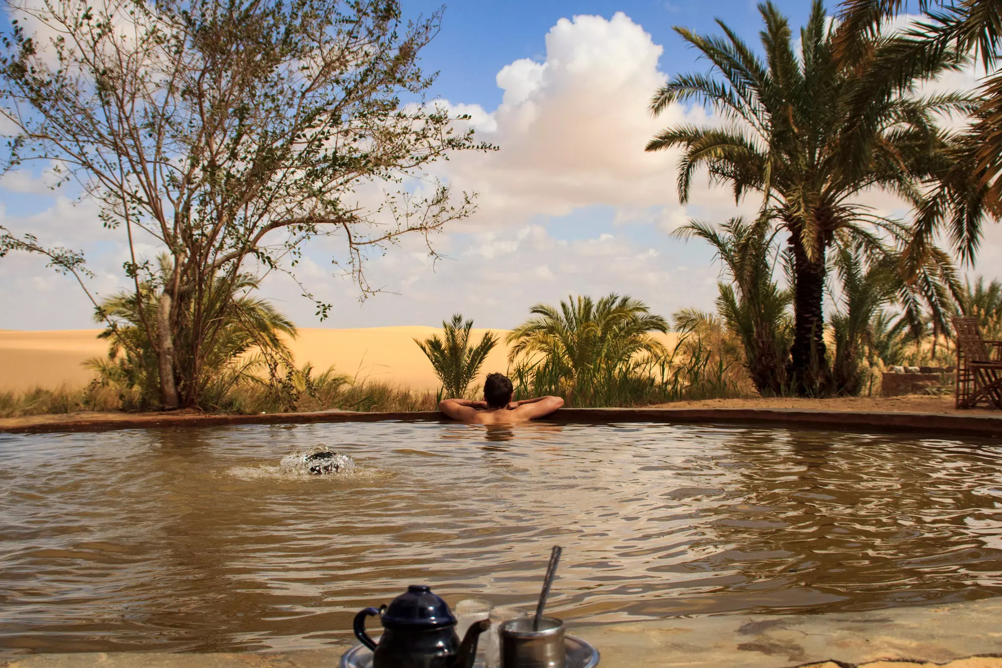 A person seen from the shoulders up in a round pool on a beach with a tea set in the foreground