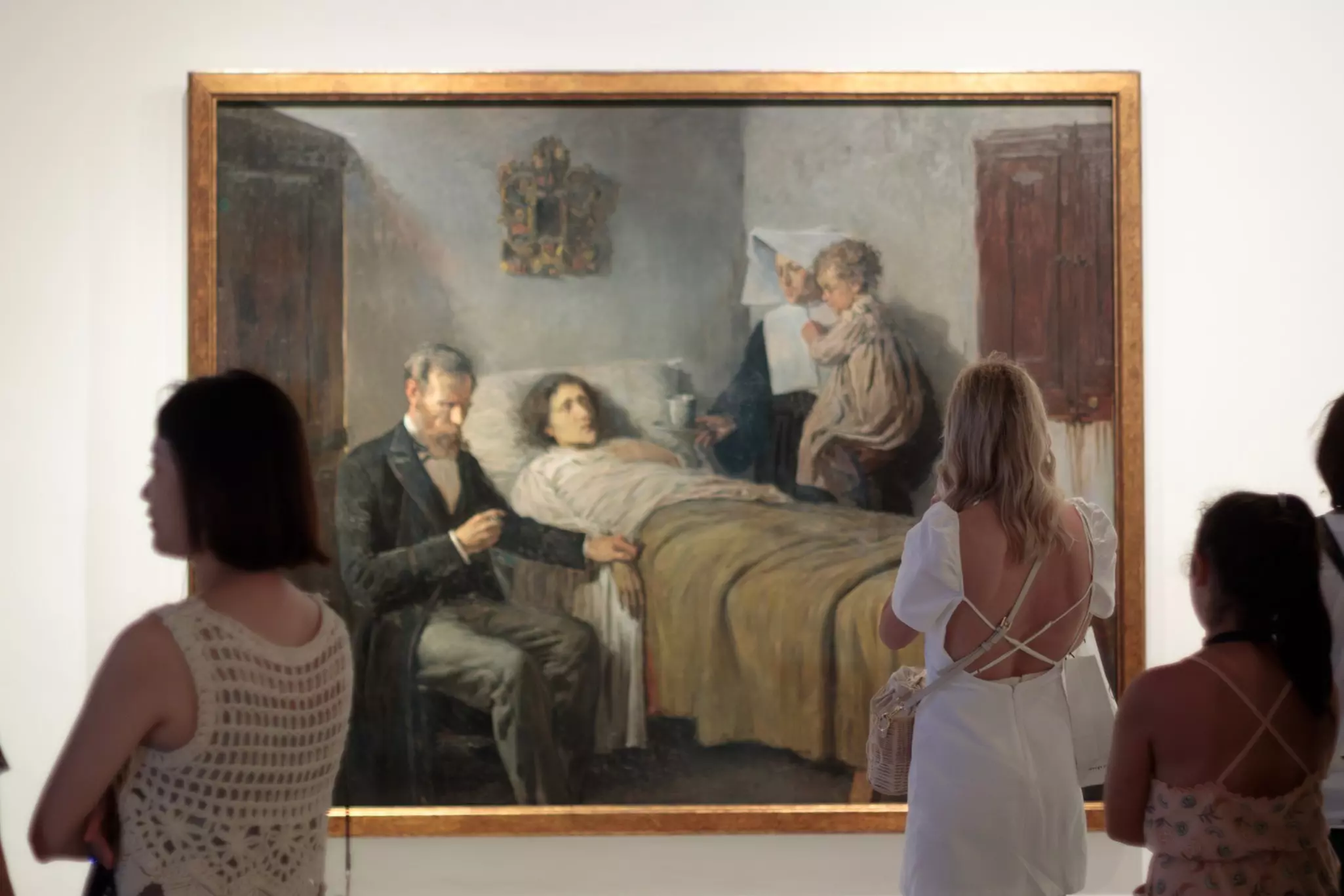 A woman in a backless white dress admires a painting of people gathered around a man who appears to be sick in bed.