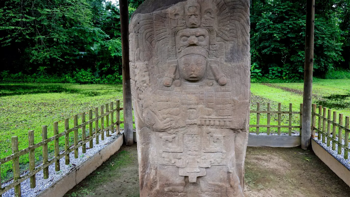 A stone stele on display outdoors behind a fence is carved with stylized faces and other motifs.