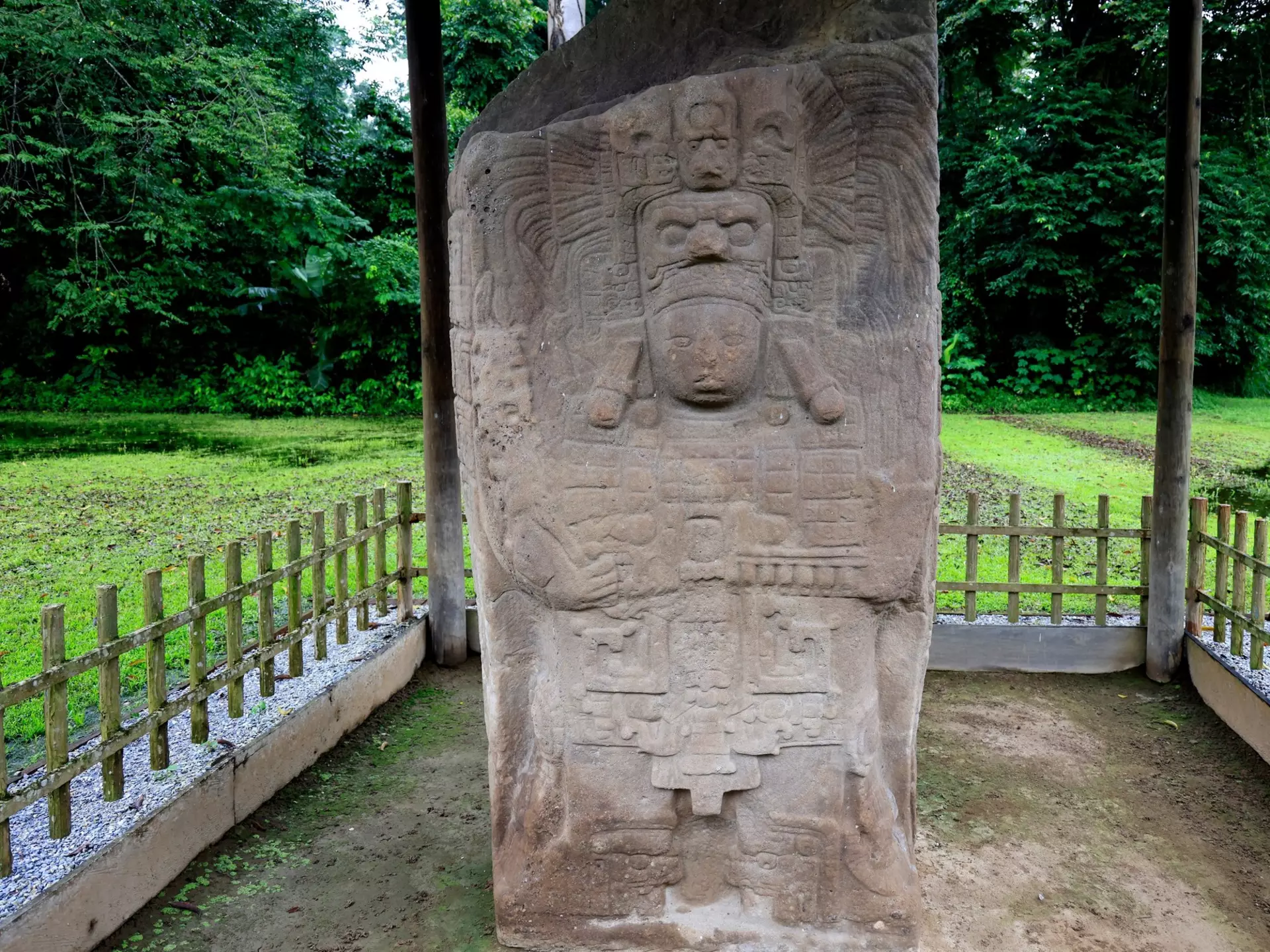 A stone stele on display outdoors behind a fence is carved with stylized faces and other motifs.