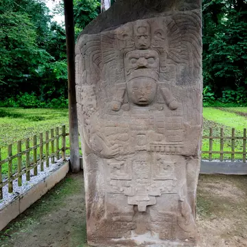 A stone stele on display outdoors behind a fence is carved with stylized faces and other motifs.