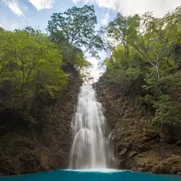 A slender waterfall pours down a dark rock gully into a turquoise pool, framed by leafy trees under a partly cloudy sky.
