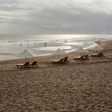 Wide sandy beach with white parasols and loungers, people walking at the water's edge under an overcast sky, Kerobokan.