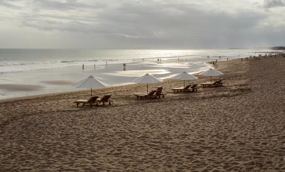 Wide sandy beach with white parasols and loungers, people walking at the water's edge under an overcast sky, Kerobokan.