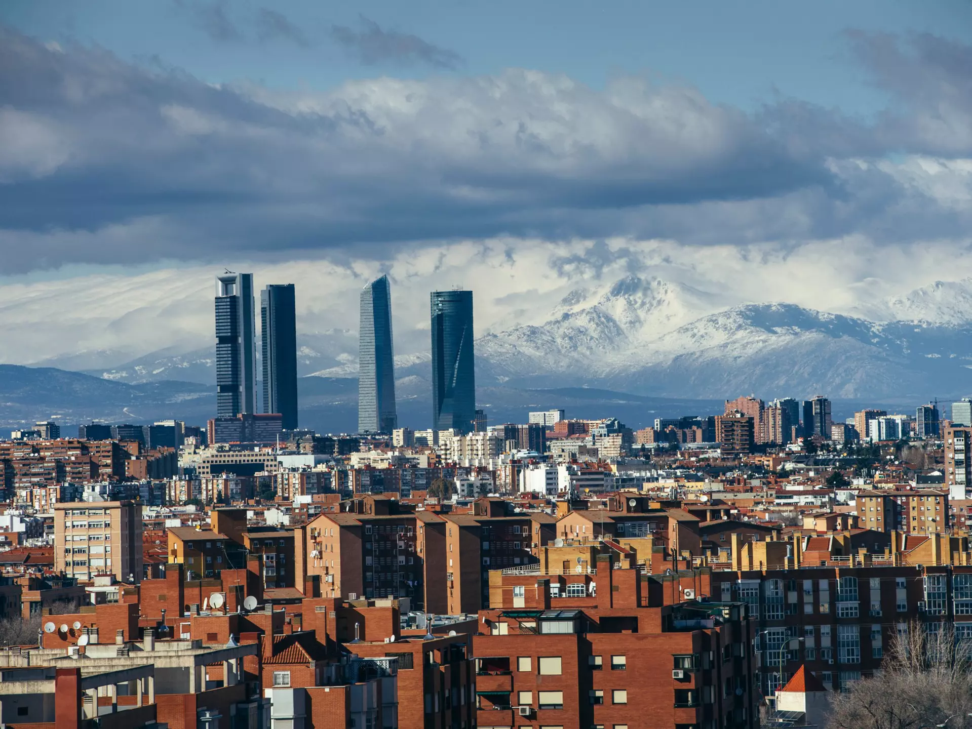 A wide view of a city skyline, including several skyscrapers, with snowy mountains in the distance.