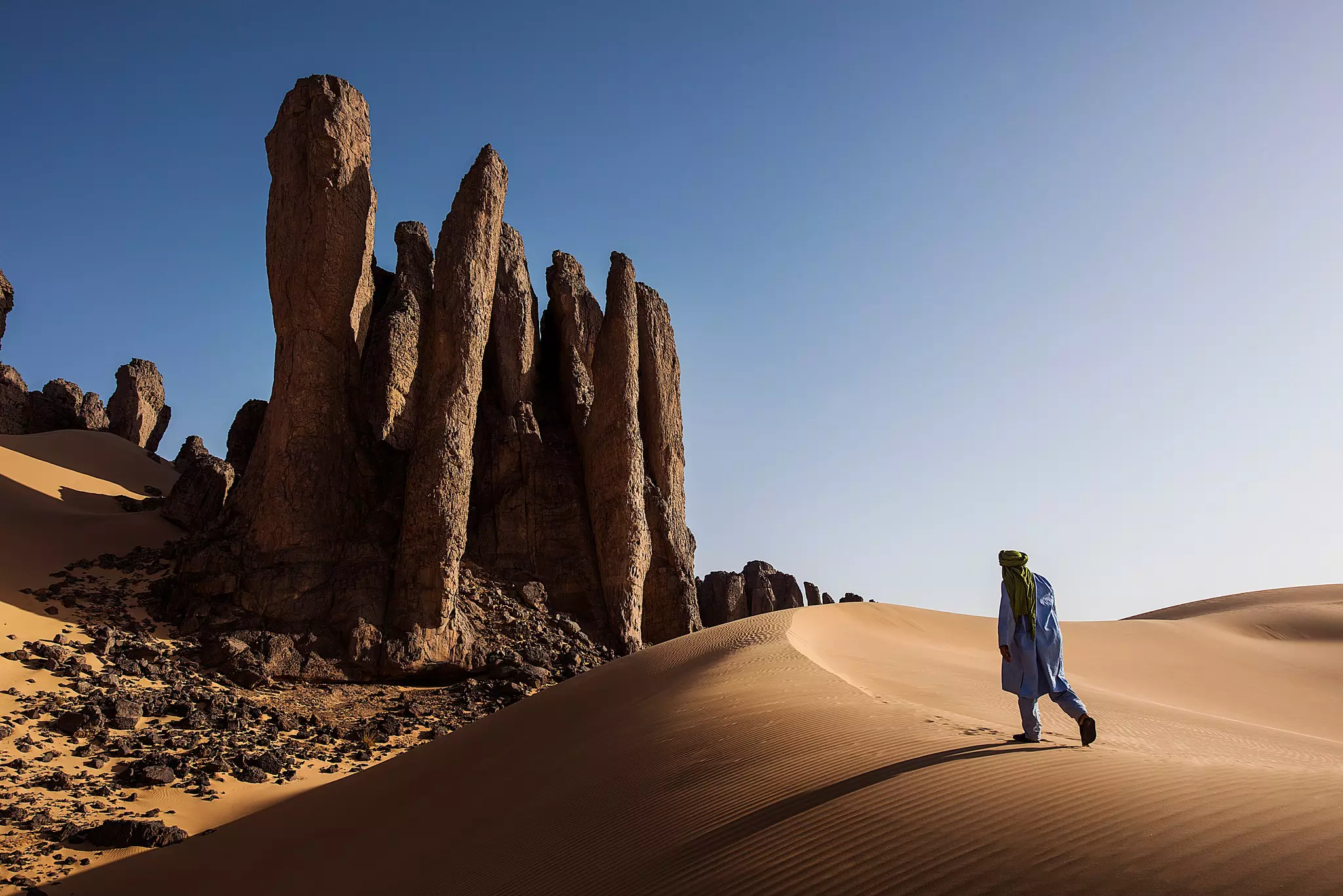 A person walking up a sand dune in Algeria toward rock formations under bright sun and blue skies.