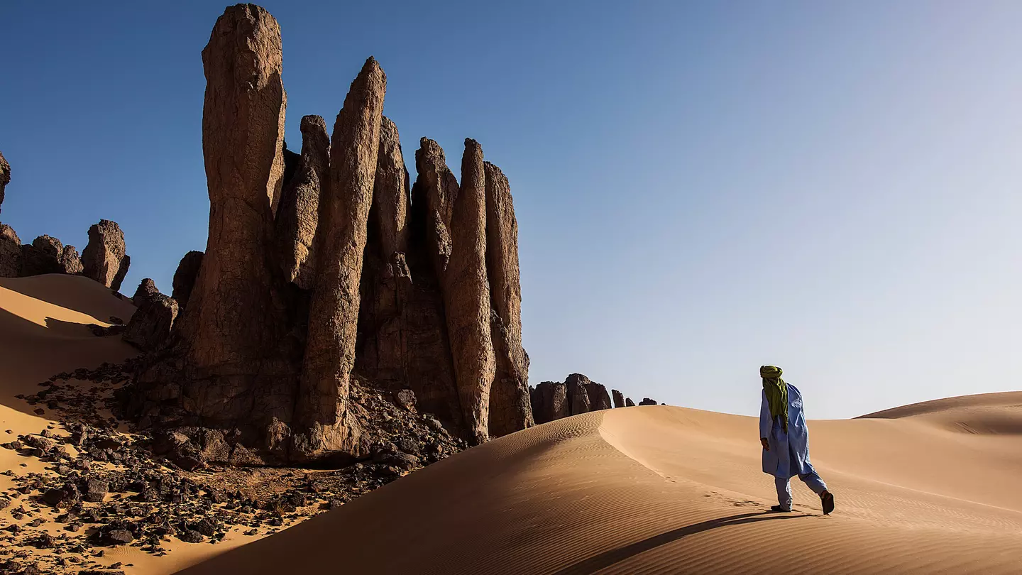 A person walking up a sand dune in Algeria toward rock formations under bright sun and blue skies.