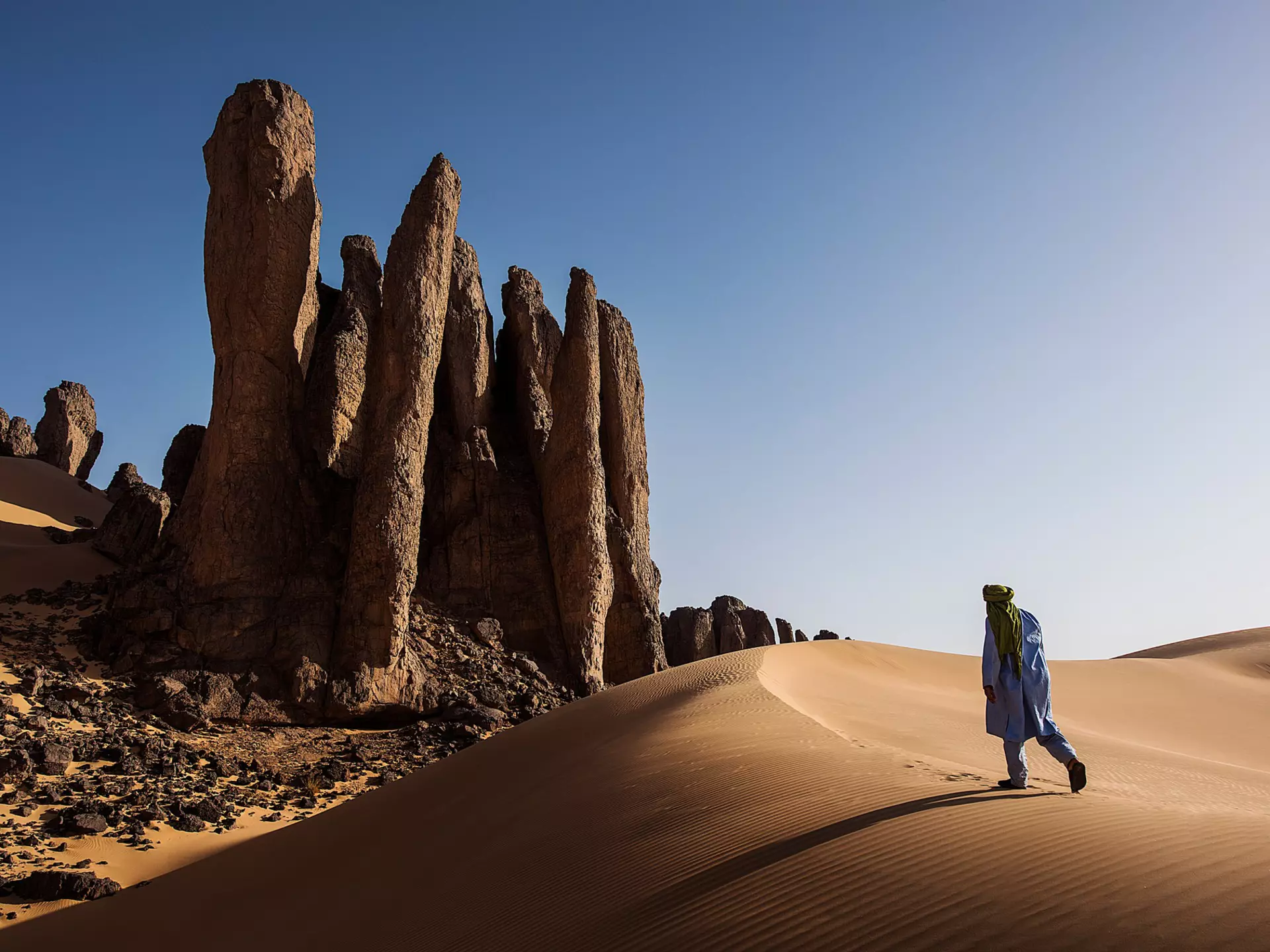 A person walking up a sand dune in Algeria toward rock formations under bright sun and blue skies.