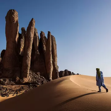A person walking up a sand dune in Algeria toward rock formations under bright sun and blue skies.