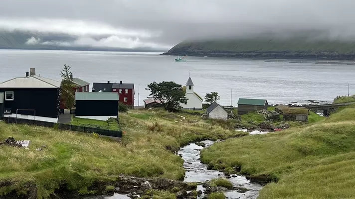 Green grass with a stream cutting through surrounded by a small church and houses with the ocean in the background