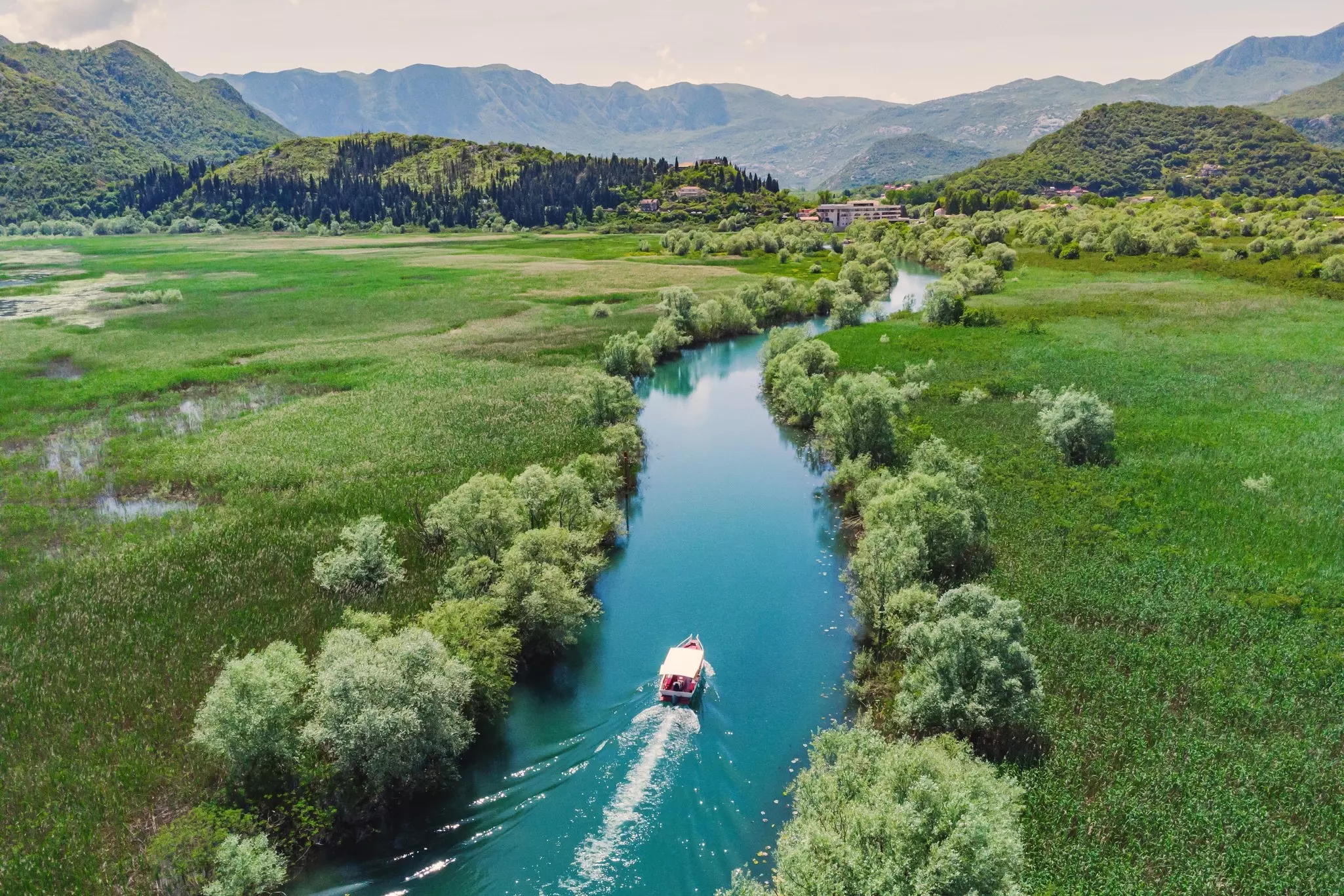 A motorboat on a bright blue waterway that is surrounded by a green wetland; there are mountains in the distance.