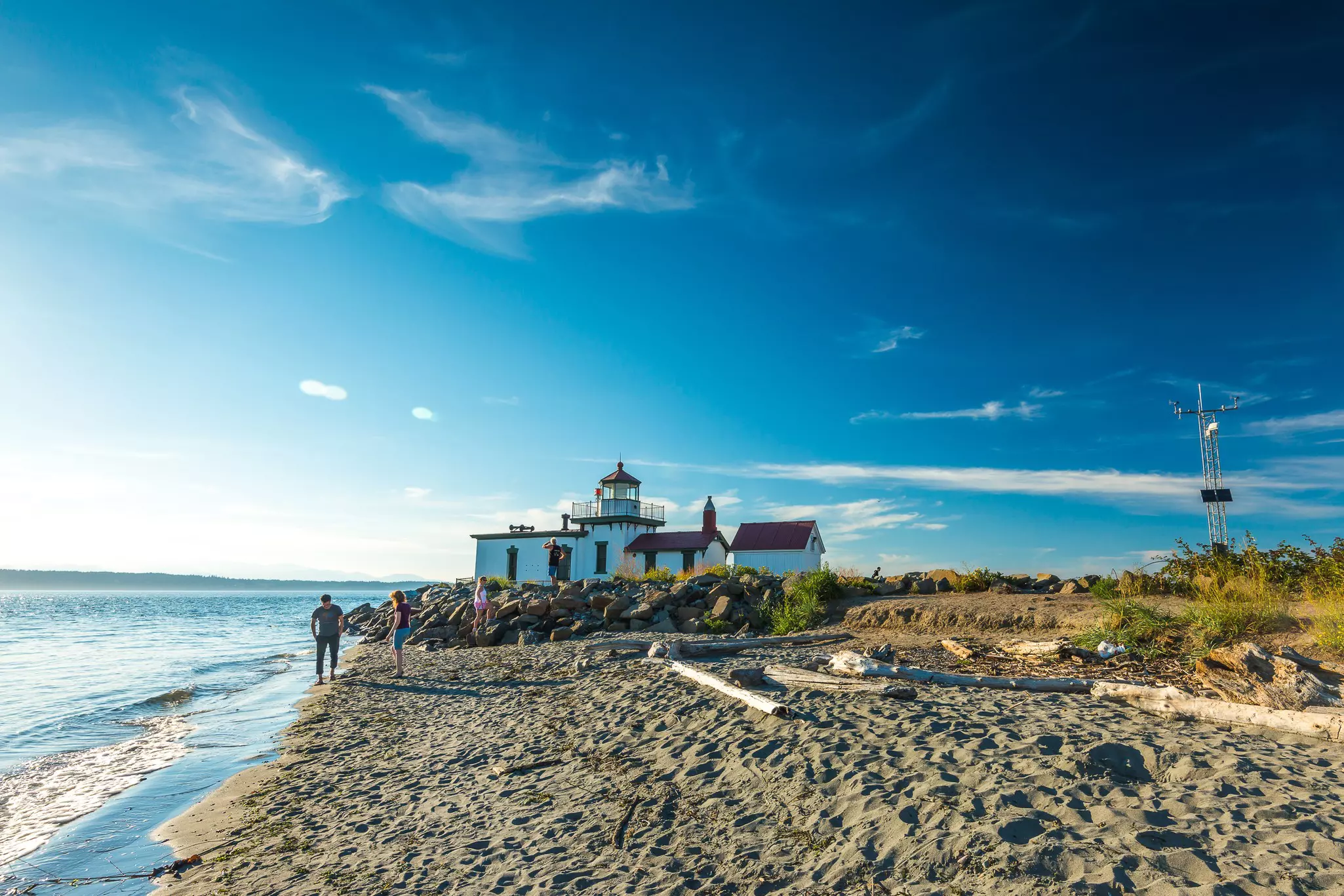 People play on the sand and clamber on a pile of boulders on a beach near a lighthouse.