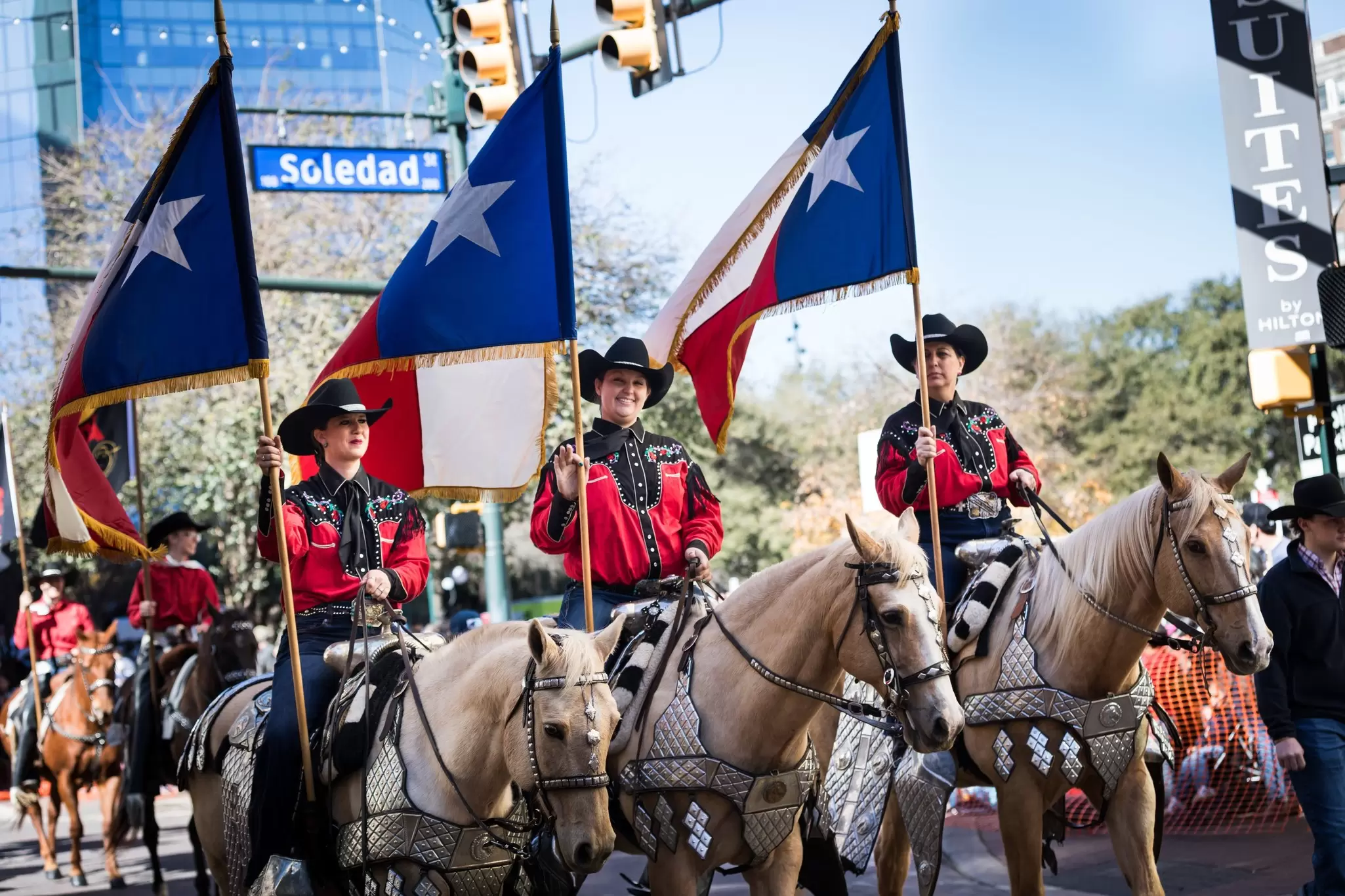 Three women on horseback wear hats and carry flags as they participate in a parde.