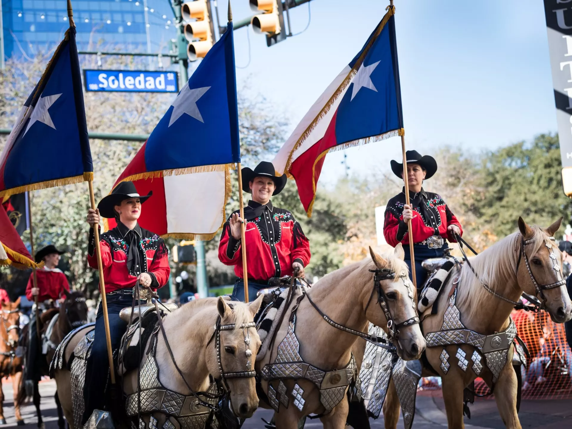 Three women on horseback wear hats and carry flags as they participate in a parde.