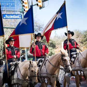 Three women on horseback wear hats and carry flags as they participate in a parde.